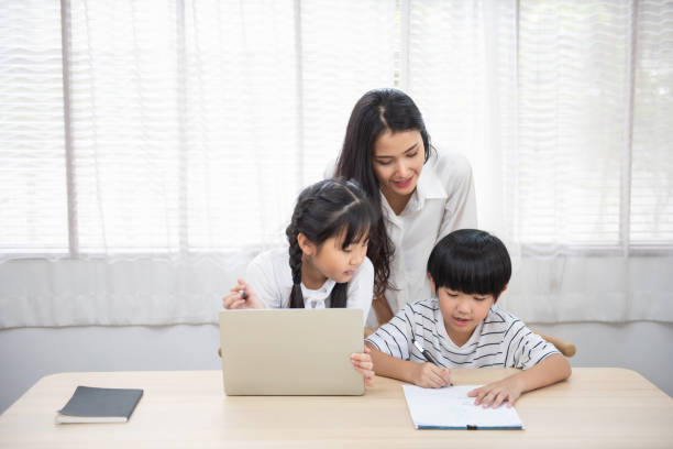 istock Asian young woman help her son do homework with daughter using laptop sit beside on table in living room at home. 1213131393