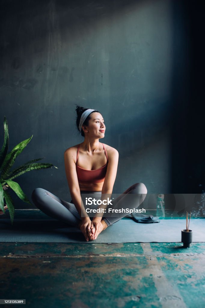 Asian Woman Sitting on an Exercise Mat and Warming Up for a Yoga Session Full-legth shot of a young Asian woman sitting on a yoga mat before exercising. Yoga Stock Photo