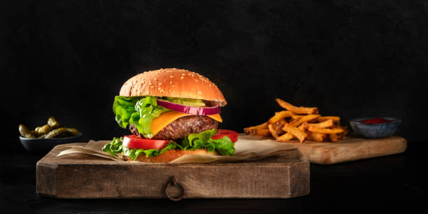 istock Burger with beef, cheese, onion, tomato, and green salad, a side view panorama on a dark background with French fries and pickles, with copy space 1213139712
