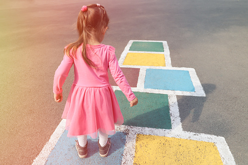 Little girl in a pink dress playing hopscotch on playground outdoors, children outdoor activities