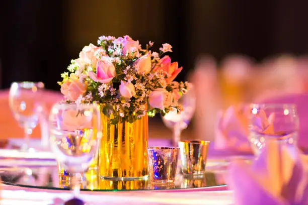 Photo of rose florist in glass vase decoration on dinner table with silverware and candle Indian wedding setup indoor with decorative lights and beautiful bokeh.