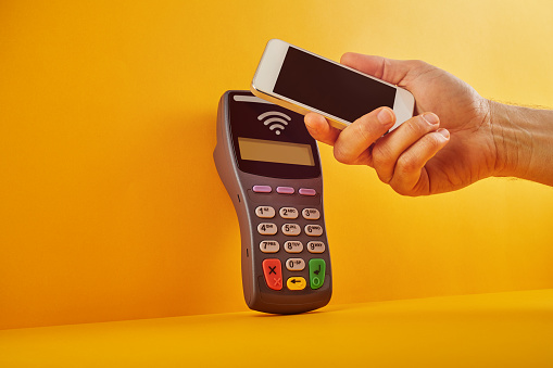 Customer paying with a smart phone via contactless NFC (Near Field Communication) machine . Electronic banking terminal over yellow background. Mobile, contactless payment concept