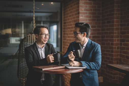 2 asian chinese white collar workers having discussion during their coffee break in lounge using laptop and digital tablet