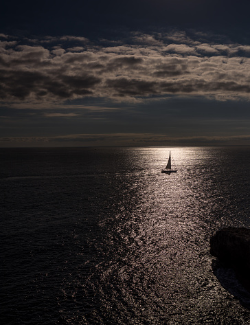 a sailboat from the coast in the Mediterranean Sea during the sunset where the water is calm, and the sailboat and sailor is on its own. the setting sun on the horizon leaves a beautiful ambience and is reflecting on the water surface