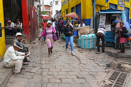 Antananarivo, Madagascar - April 24, 2019: Two unknown young Malagasy girls walking cobblestone road, more people around at their stalls, or sitting on ground - typical scene in streets of Tana city