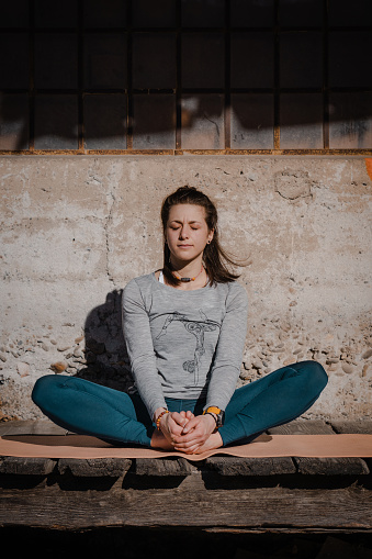 Young yoga woman sitting cross-legged on a wooden walkway in front of an old factory. She warms up for her yoga session, which is part of her daily routine, workout.
