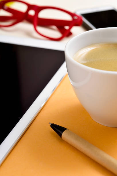 closeup of an office or home office desk full of things, such as a cup with white coffee, a pen, a tablet, a smartphone and a pair of red eyeglasses