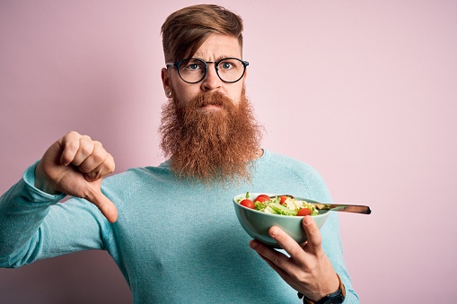 Redhead Irish healthy man with beard eating vegetarian green salad over pink background with angry face, negative sign showing dislike with thumbs down, rejection concept