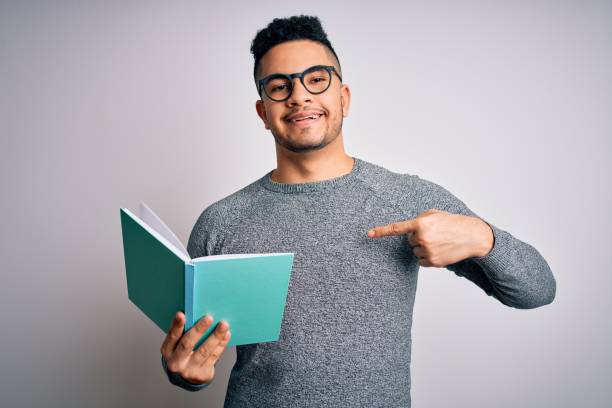 Young handsome smart student man reading book over isolated white background with surprise face pointing finger to himself