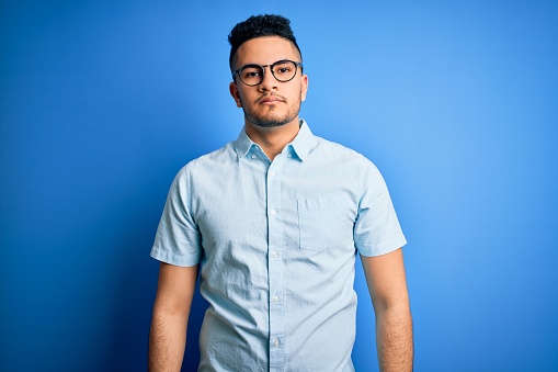 Young handsome man wearing casual summer shirt and glasses over isolated blue background with serious expression on face. Simple and natural looking at the camera.