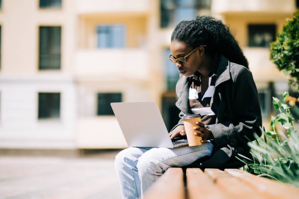 istock Pretty African American girl sitting on bench and talking on cellphone while working on laptop sitting at bench with coffee on street 1213211609