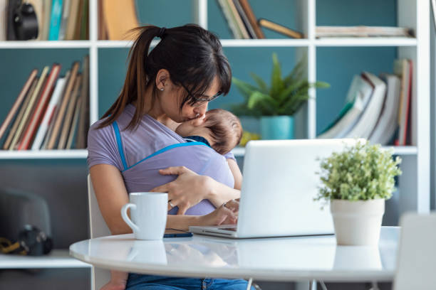 istock Pretty young mother kissing her baby in sling while working with laptop at home. 1213212708