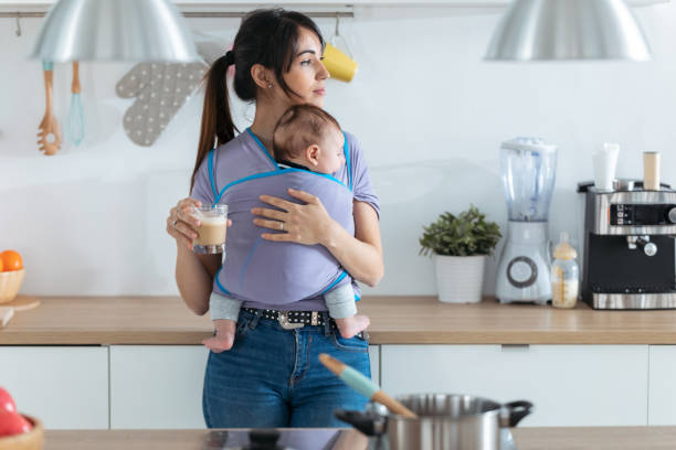 bella giovane madre con il bambino piccolo in fionda a bere caffè in cucina a casa. - carry clothes foto e immagini stock