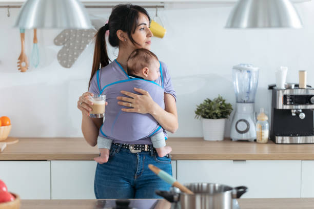 Pretty young mother with little baby in sling drinking coffee in the kitchen at home. Shot of pretty young mother with little baby in sling drinking coffee in the kitchen at home. baby carrier stock pictures, royalty-free photos & images