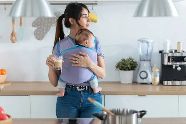 Shot of pretty young mother with little baby in sling drinking coffee in the kitchen at home.