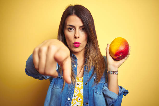 Young beautiful woman eating fresh healthy mango over yellow background pointing with finger to the camera and to you, hand sign, positive and confident gesture from the front
