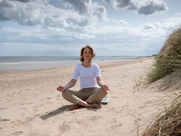 Mature woman meditating in Lotus Pose on the beach under a dramatic sky. Knokke, Belgium.