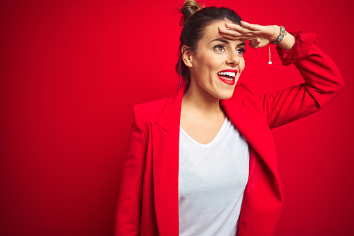 Young beautiful business woman standing over red isolated background very happy and smiling looking far away with hand over head. Searching concept.