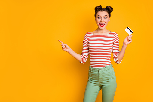 Photo of pretty excited funny lady hold credit card wireless online money, direct finger empty space wear striped red white shirt green pants isolated bright yellow color background