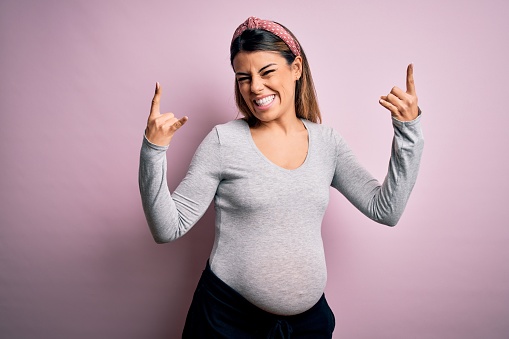 Young beautiful brunette woman pregnant expecting baby over isolated pink background shouting with crazy expression doing rock symbol with hands up. Music star. Heavy concept.