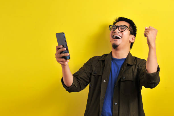 Asian man holding smartphone with winning gesture Asian man holding smartphone with winning gesture. Asian bussinesman winning gift or lottery, on yellow background playing card photos stock pictures, royalty-free photos & images