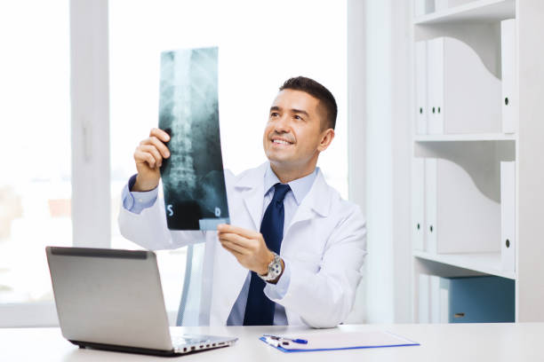 smiling male doctor in white coat looking at x-ray healthcare, technology, rontgen, people and medicine concept - smiling male doctor in white coat with laptop computer looking at x-ray in medical office orthopedist stock pictures, royalty-free photos & images