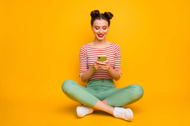 Full length photo of pretty attractive lady sit floor legs crossed hold telephone typing new post blog text wear striped red white shirt green pants shoes isolated yellow color background Full length photo of pretty attractive lady sit floor legs crossed hold telephone, typing new post blog text wear striped red white shirt green pants shoes isolated yellow color background topknot stock pictures, royalty-free photos & images