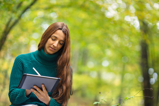 A woman in her 30s outdoors in nature, working on a digital tablet with a digital pen.