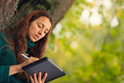 A woman sits concentrated by a tree outdoors, using a tablet computer and digital pen.