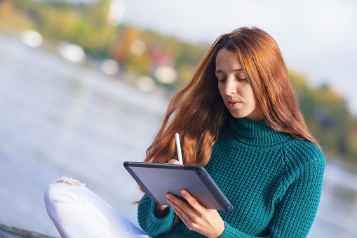 A woman sits by the edge of a lake, busy with a digital tablet and pen.