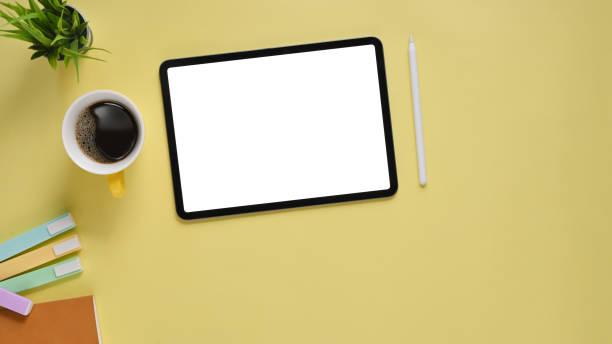 istock Top view image of yellow color working table. Flat lay cropped black computer tablet with white blank screen, coffee cup, notebook, potted plant and marker pen. Colorful workplace concept. 1213243108