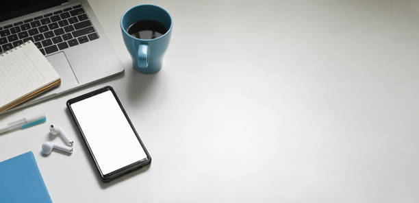 Top view image of working table with accessories putting on it. White blank screen smartphone, coffee cup, wireless earphone, notebook, pen and computer laptop flat lay on white space.