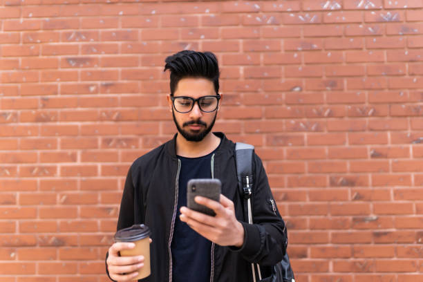 istock Young handsome indian student man with phone and coffee cup standing against brick wall 1213249436