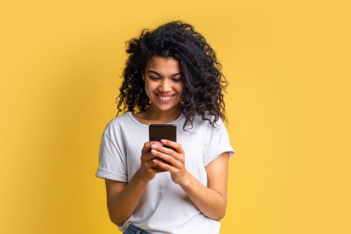 Cute happy blogger girl with perfect smile in basic white t-shirt using her mobile phone answering to her followers, texting to her boyfriend, browsing various applcations. Device addiction concept.