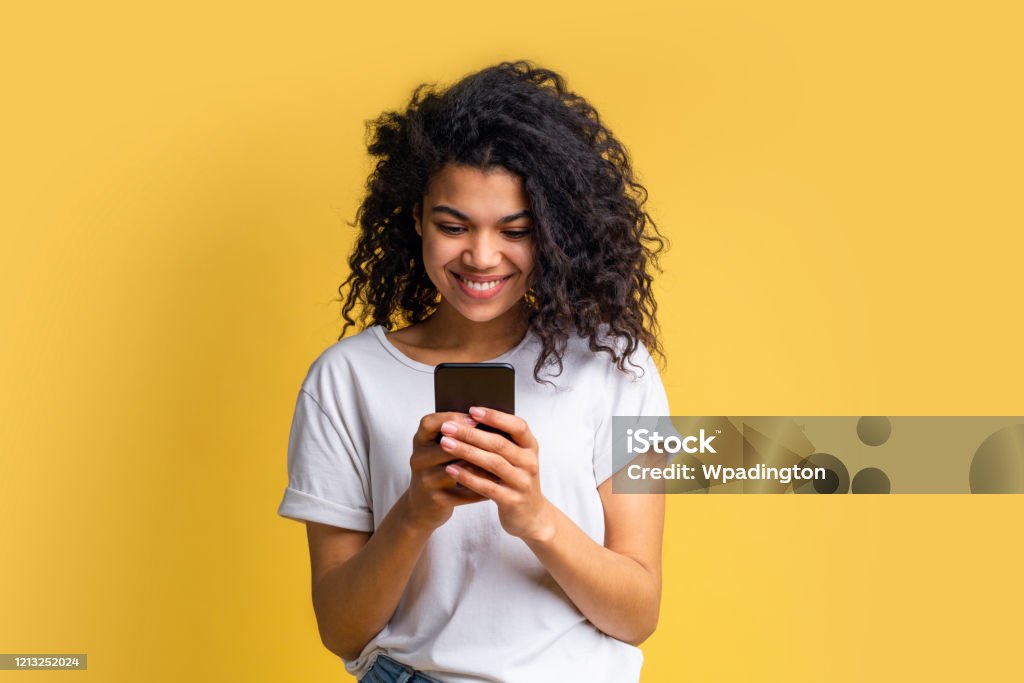 Portrait of attractive young african american girl using mobile phone Cute happy blogger girl with perfect smile in basic white t-shirt using her mobile phone answering to her followers, texting to her boyfriend, browsing various applcations. Device addiction concept. Mobile Phone Stock Photo