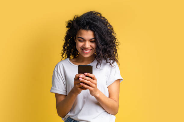 Portrait of attractive young african american girl using mobile phone Cute happy blogger girl with perfect smile in basic white t-shirt using her mobile phone answering to her followers, texting to her boyfriend, browsing various applcations. Device addiction concept. translation photos stock pictures, royalty-free photos & images