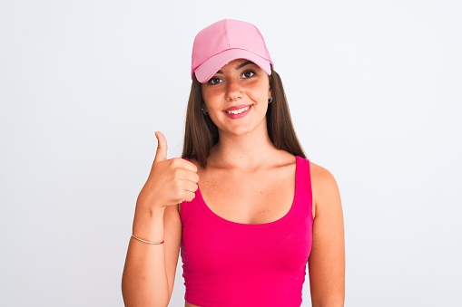 Young beautiful girl wearing pink casual t-shirt and cap over isolated white background doing happy thumbs up gesture with hand. Approving expression looking at the camera with showing success.