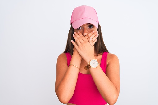 Young beautiful girl wearing pink casual t-shirt and cap over isolated white background shocked covering mouth with hands for mistake. Secret concept.