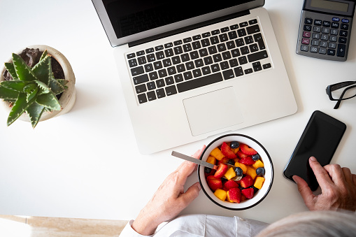 Above view of a colorful fruit salad on the desk - laptop and cellphone on the table - elderly woman taking a break