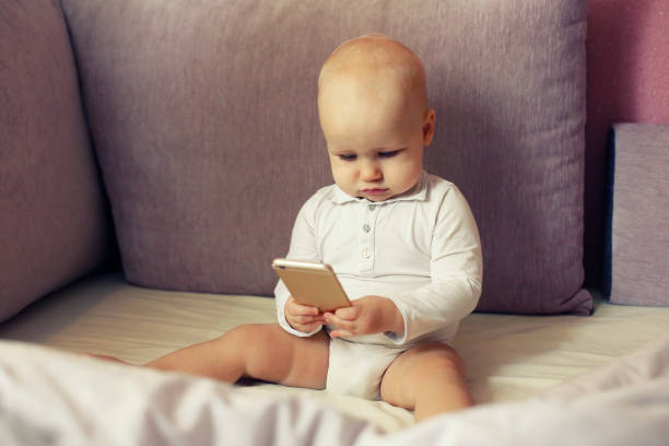 Baby infant in white sit on sofa bed and look at mobile smartphone screen, new generation and electronics stock photo