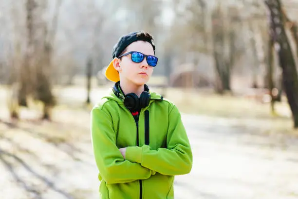 March 18, 2020 - Kabaty, Poland: cute serious intelligent teen boy standing in a sunny park wearing sunglasses, chin up