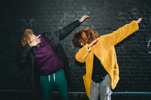 Two young adults making dabbing movement against a black bricks wall. They are wearing hip and colorful clothes.