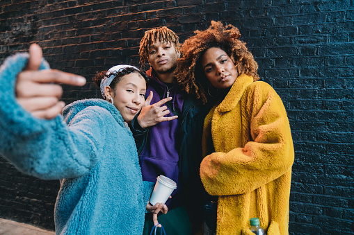 Portrait of three friends against a black bricks wall, making gestures in front of the camera. They are wearing colorful and hip clothes.