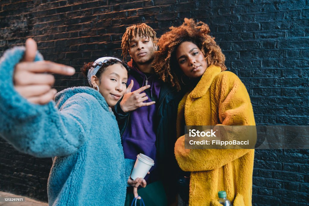 Portrait of three friends against a black bricks wall, making gestures in front of the camera Portrait of three friends against a black bricks wall, making gestures in front of the camera. They are wearing colorful and hip clothes. Teenager Stock Photo