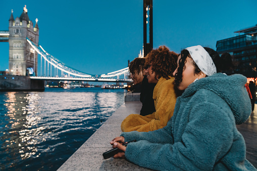 Three friends looking at the view near Tower Bridge in London. Night time. They are wearing baggy and colorful clothes.
