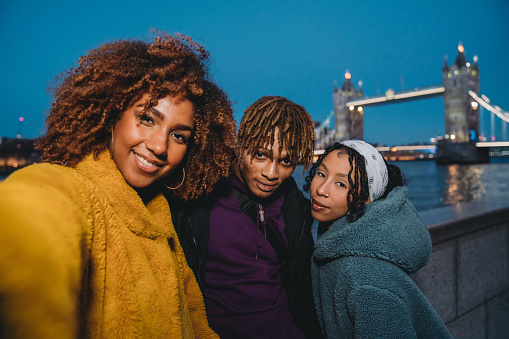 Pov view of three young adult hip friends taking a selfie in London near Tower Bridge. Night time. They are wearing baggy hip and colorful clothes.