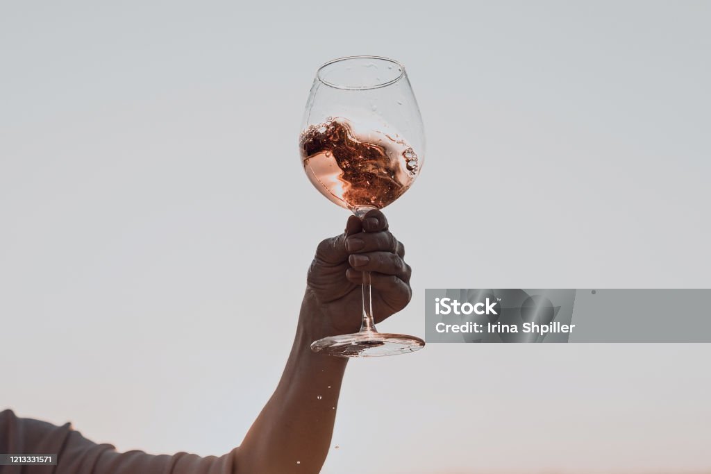 Glass of wine with splashes in woman's hand against the sunset sky. Glass of wine with splashes in woman's hand against the sunset sky. Cropped. Wine Stock Photo