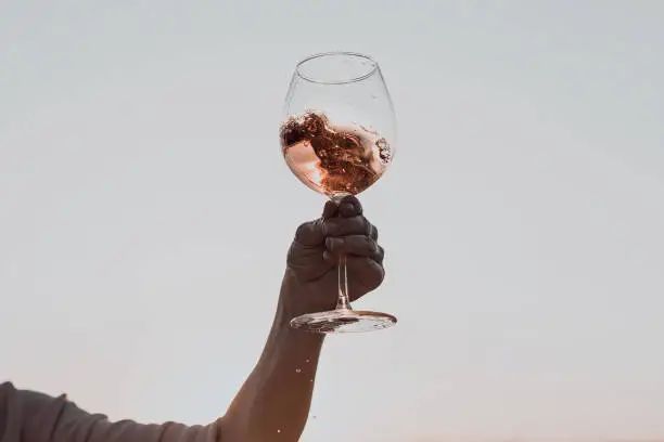 Photo of Glass of wine with splashes in woman's hand against the sunset sky.