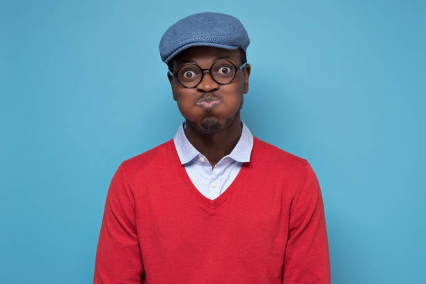 Young african man in red sweater and blue hat puffing cheeks holding breath Handsome young african man in red sweater and blue hat puffing cheeks holding breath isolated over blue wall. Studio shot puffed up cheeks stock pictures, royalty-free photos & images
