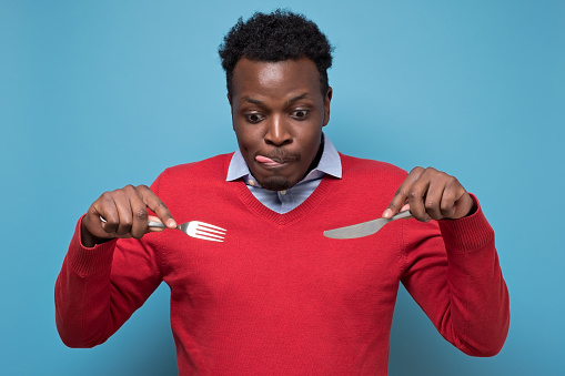 Young hungry crazy african american man in red sweater holding a fork and a knife. Isolated on blue background